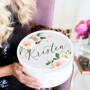 May include: White round gift box with a floral design and the name "Kristen" written in black script. The flowers are pink, white, and green.