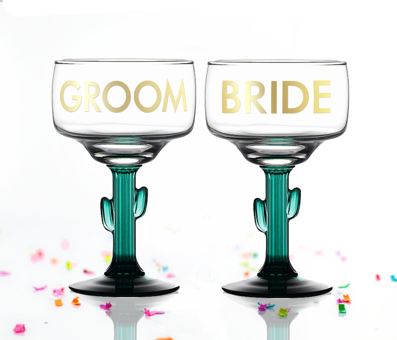 May include: Two clear glass margarita glasses with green cactus stems. The glasses are decorated with gold lettering, one says "Groom" and the other says "Bride".