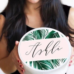 May include: White round gift box with a pink and green tropical leaf design. The box has the name "Ashli" written in black cursive on a pink band.