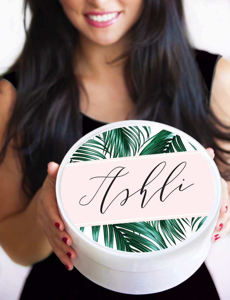 May include: White round gift box with a pink and green tropical leaf design. The box has the name "Ashli" written in black cursive on a pink band.
