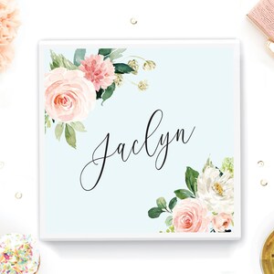 May include: A light blue square box with a white border. The box has a floral design with pink and white flowers and green leaves. The name "Jaclyn" is written in black cursive script in the center of the box.