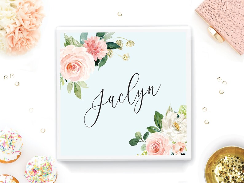 May include: A light blue square box with a white border. The box has a floral design with pink and white flowers and green leaves. The name "Jaclyn" is written in black cursive script in the center of the box.