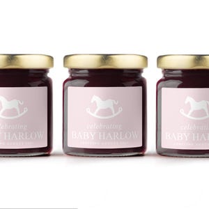 May include: Three glass jars with gold lids filled with a dark red preserve. Each jar has a pink label with a white rocking horse graphic and the text "celebrating BABY HARLOW ARRIVING AUGUST 2023".