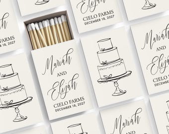 May include: A collection of rectangular matchboxes with a cream background, featuring a black line drawing of a three-tiered wedding cake on a pedestal. The text reads Mariah and Elijah, Cielo Farms, December 18, 2027. One box is open, showing wooden matches with white tips.