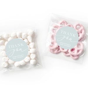 Peut inclure: Deux sacs en plastique transparents remplis de cadeaux de f&ecirc;te. Un sac contient des guimauves blanches et l'autre des bretzels roses. Les deux sacs ont un autocollant rond bleu clair avec le texte "Merci" en &eacute;criture blanche et "Baby Shower de Samantha" en texte blanc.