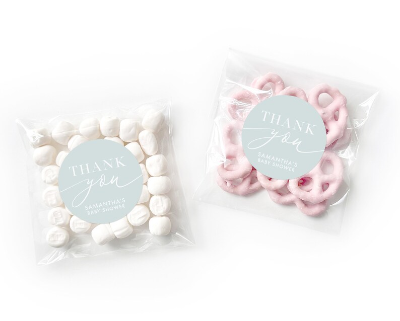 Peut inclure: Deux sacs en plastique transparents remplis de cadeaux de f&ecirc;te. Un sac contient des guimauves blanches et l'autre des bretzels roses. Les deux sacs ont un autocollant rond bleu clair avec le texte "Merci" en &eacute;criture blanche et "Baby Shower de Samantha" en texte blanc.