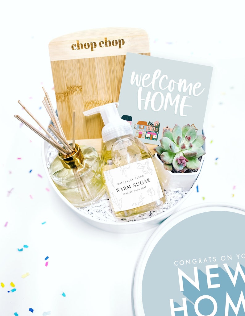 Puede incluir: Una caja de regalo blanca con una tapa azul. La caja contiene una tabla de cortar de madera con el texto "chop chop" grabado en ella, una botella de jab&oacute;n de manos, una peque&ntilde;a suculenta en maceta y una tarjeta que dice "bienvenido a casa".