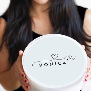 Puede incluir: Una caja redonda blanca con una tapa azul claro. La tapa tiene un logotipo negro en cursiva que dice "&hearts;m MONICA".
