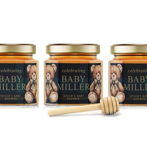 May include: Three small glass jars filled with honey, each with a label that reads "celebrating Baby Miller, Taylor's Baby Shower". The labels feature a cartoon teddy bear with a bow tie and a plaid background. A wooden honey dipper is in the foreground.