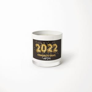 May include: A white glass candle jar featuring a black rectangular label with gold foil-style text that reads 2022, CONGRATS GRAD, and the name Sabrina, accented by small gold dots resembling confetti against a dark background.
