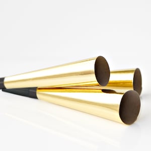May include: Three gold party noisemakers, or blowers, are stacked on top of each other. The noisemakers are made of cardboard and have a black stripe at the bottom.