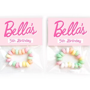 May include: Two clear plastic bags each containing a candy bracelet. Each bag has a pink card with the name "Bella's" in cursive and "5th Birthday" in a smaller font. The candy bracelets are made of pastel-colored candies.