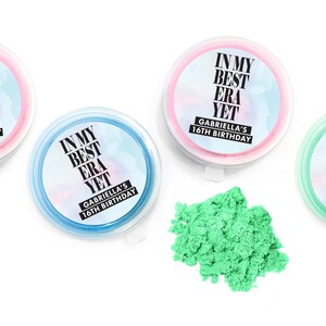 May include: Three clear plastic containers with lids, each filled with a different color of sand. The containers have labels that say "In My Best Era Yet" and "Gabriella's 16th Birthday".