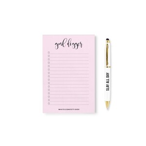 May include: A pink notepad with the text "goal digger" and a checklist. A white pen with gold accents and the text "SLAY ALL DAY" is next to the notepad.