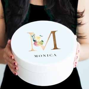 K&ouml;nnte beinhalten: Wei&szlig;e runde Geschenkbox mit einem goldenen Buchstaben "M", verziert mit Aquarellblumen und dem darunter gedruckten Namen "MONICA".
