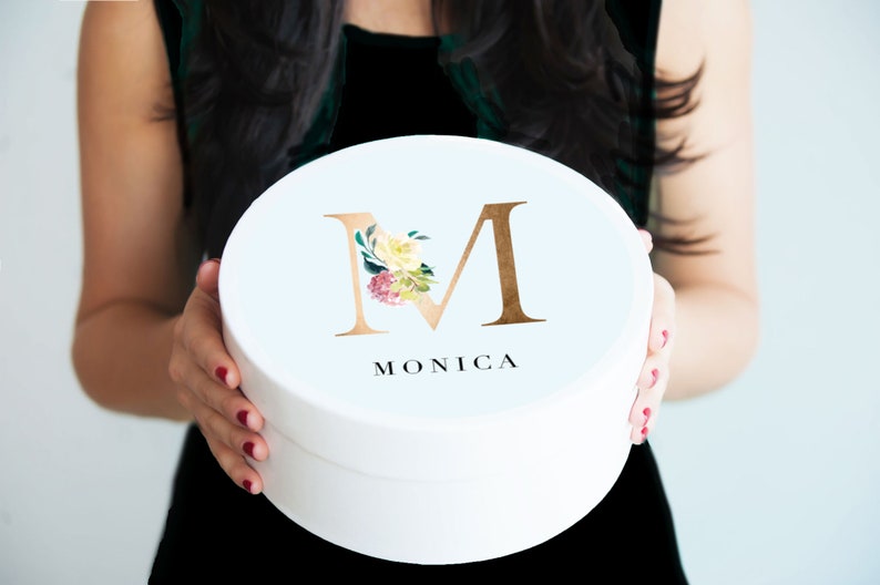 K&ouml;nnte beinhalten: Wei&szlig;e runde Geschenkbox mit einem goldenen Buchstaben "M", verziert mit Aquarellblumen und dem darunter gedruckten Namen "MONICA".