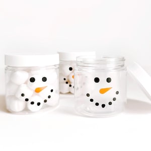 May include: Three clear plastic jars with white screw-on lids. Each jar features a snowman face graphic printed on the front, consisting of two black circular eyes, an orange carrot-shaped nose, and a curved mouth made of small black dots.