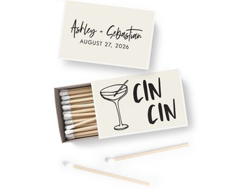 May include: A white matchbox with a black line drawing of a martini glass and the text "CIN CIN" in black. The top of the matchbox has a beige label with the text "Ashley + Sebastian AUGUST 27, 2026" in black.