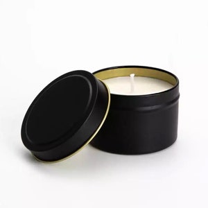 May include: A black candle tin with a gold rim and a white candle inside. The lid is off and sitting next to the tin.