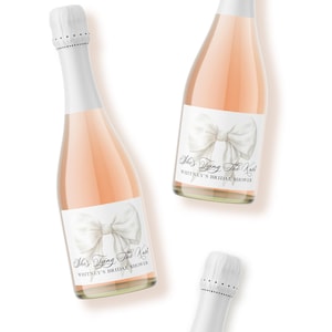 May include: Two glass bottles filled with a light pink liquid, each featuring a white label with a large illustration of a white bow. The text on the labels reads "She's Tying The Knot" and "WHITNEY'S BRIDAL SHOWER" in black script and serif fonts.