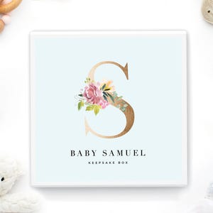 May include: A square white keepsake box with a light blue background featuring a large gold letter S decorated with watercolor-style pink and purple flowers and green leaves. The text BABY SAMUEL KEEPSAKE BOX is printed in black below the letter.