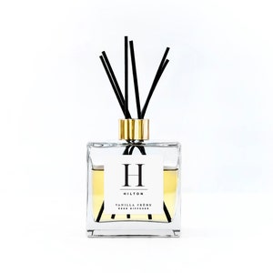 May include: A clear glass bottle with a gold lid and black reeds. The bottle contains a yellow liquid and has a white label with the text "HILTON" and "VANILLA CREME REED DIFFUSER".