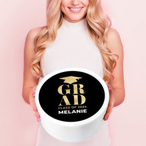 May include: A white round cake box with a black circle on top. The circle has gold text that says "GRAD" with a graduation cap above the word. Below the word "GRAD" is the text "CLASS OF 2024 MELANIE".