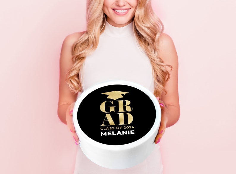 May include: A white round cake box with a black circle on top. The circle has gold text that says "GRAD" with a graduation cap above the word. Below the word "GRAD" is the text "CLASS OF 2024 MELANIE".