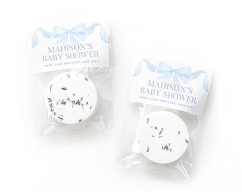 May include: Two round white bath bombs with visible dried botanical pieces, each packaged in a clear plastic bag with a white header card featuring a light blue watercolor bow and the text: MADISON'S BABY SHOWER BABY LIAM ARRIVING JUNE 2027.