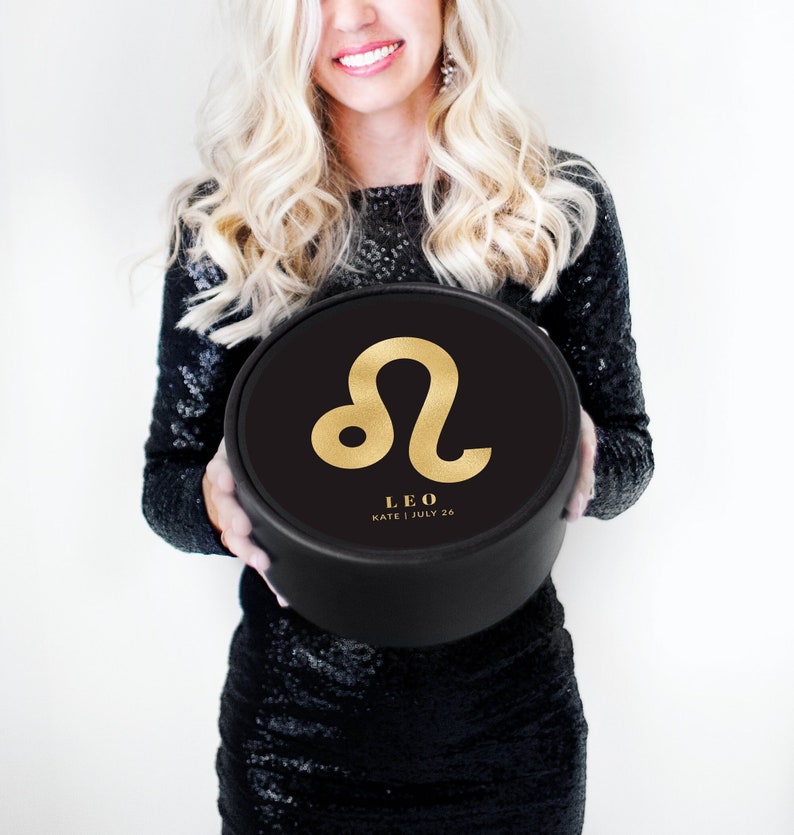 K&ouml;nnte beinhalten: Eine schwarze runde Box mit einem goldenen L&ouml;we-Sternzeichen und dem Text "LEO, KATE | JULY 26".