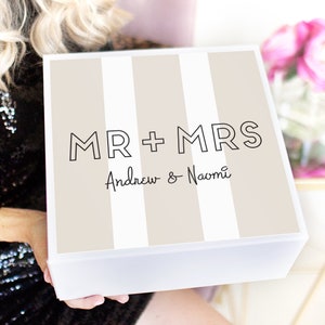 May include: A white gift box with a beige and white striped pattern. The box has the text "MR + MRS Andrew & Naomi" printed on the front.