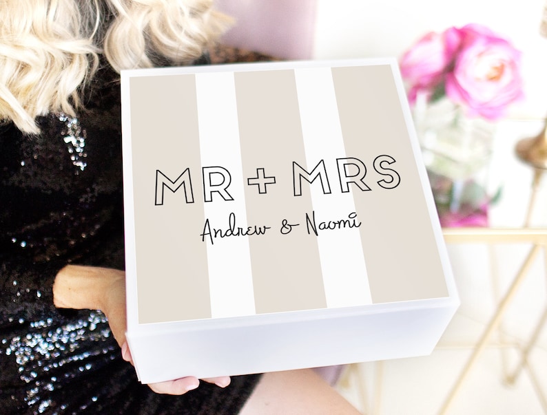 May include: A white gift box with a beige and white striped pattern. The box has the text "MR + MRS Andrew & Naomi" printed on the front.