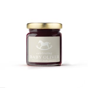 May include: A small glass jar with a gold lid filled with a dark red preserve. The jar has a label with a rocking horse illustration and the text "celebrating BABY OLIVER VICTORIA'S BABY SHOWER".
