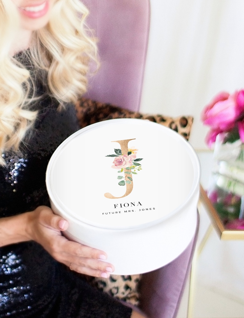 May include: White round gift box with a gold letter J decorated with pink and green floral accents. The box has the name Fiona and the text "Future Mrs. Jones" printed on it.