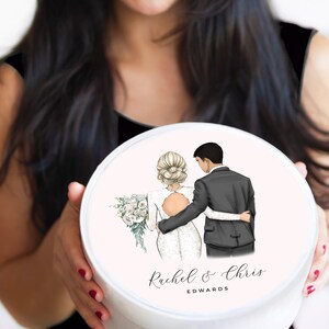 May include: White round box with a pink lid featuring a cartoon illustration of a bride and groom. The bride is wearing a white dress and the groom is wearing a black suit. The text "Rachel & Chris EDWARDS" is printed on the lid.