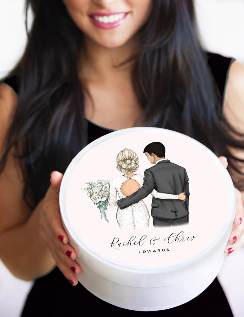 May include: White round box with a pink lid featuring a cartoon illustration of a bride and groom. The bride is wearing a white dress and the groom is wearing a black suit. The text "Rachel & Chris EDWARDS" is printed on the lid.