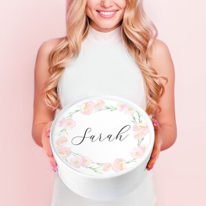 May include: A white round gift box with a pink floral wreath design and the name "Sarah" written in black calligraphy.