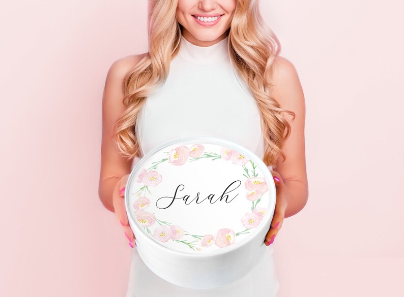 May include: A white round gift box with a pink floral wreath design and the name "Sarah" written in black calligraphy.