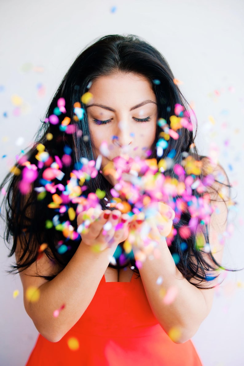 Pu&ograve; includere: Una donna con lunghi capelli neri soffia coriandoli dalle sue mani. I coriandoli sono multicolori e creano un'atmosfera festosa.