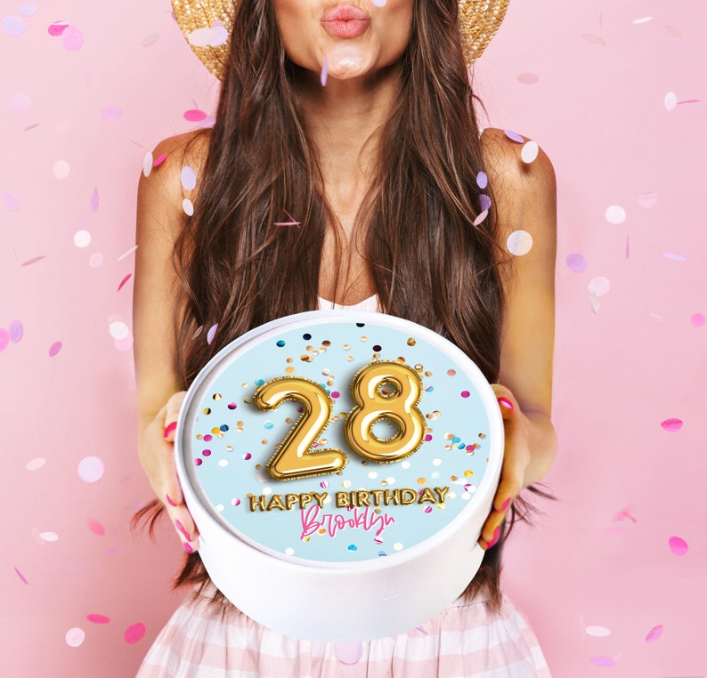 May include: A white round cake box with a blue background and gold foil number 28 balloons. The text "Happy Birthday Brooklyn" is written in pink and white on the box.