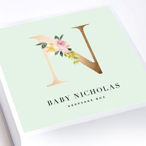 May include: A white keepsake box with a light green top featuring a large metallic gold letter N decorated with a watercolor pink rose and green leaves. The text BABY NICHOLAS KEEPSAKE BOX is printed in black below the letter N on the box lid.