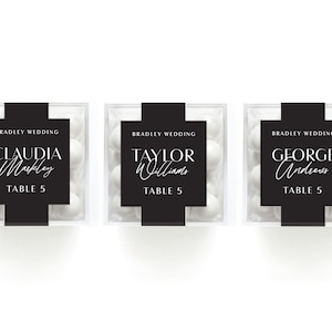 May include: Three clear boxes filled with white candy, each with a black label that says "Bradley Wedding" and the name of a guest, "Claudia Markley", "Taylor Williams", and "George Andrews", followed by "Table 5".