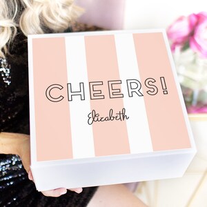 May include: A white gift box with a pink and white striped design. The box has the text "CHEERS!" in black and the name "Elizabeth" written in cursive.