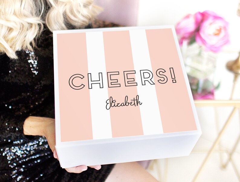 May include: A white gift box with a pink and white striped design. The box has the text "CHEERS!" in black and the name "Elizabeth" written in cursive.