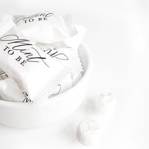 May include: A white ceramic bowl filled with white rectangular candy wrappers featuring the black printed text Mint TO BE. Two additional white wrapped candies are positioned outside the bowl on a bright white surface.