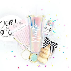 May include: A gift box filled with items for a birthday or special occasion. The box includes a pink and white iridescent tumbler with a gold lid and straw, a small jar of colorful sprinkles, a pink and white notepad with the text "Let's do this", a small candle, and a bag of cookies. The tumbler and sprinkle jar are personalized with the name "Nicole".