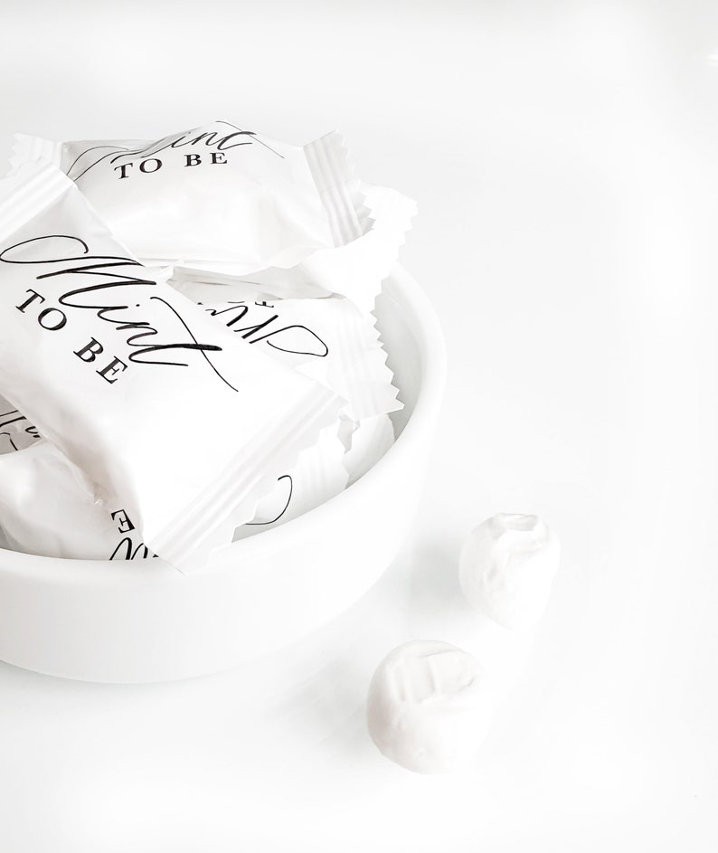 May include: White favor bags with black text that reads "Mint to be" in a bowl. Two white mints are on a white surface.