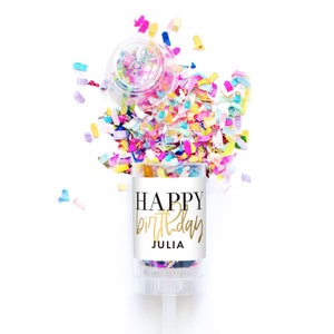 May include: A white confetti cannon with a label that says "HAPPY birthday JULIA" in gold lettering. The cannon is surrounded by colorful confetti.