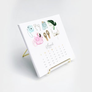 May include: A white desk calendar with a gold stand. The calendar features a watercolor illustration of summer accessories, including a flamingo float, sandals, a camera, and a swimsuit. The month of August is highlighted in gold lettering.