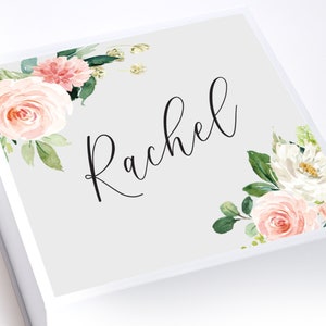 May include: A white gift box with a gray lid. The lid is decorated with a floral design in shades of pink and white. The name "Rachel" is written in black cursive script on the lid.